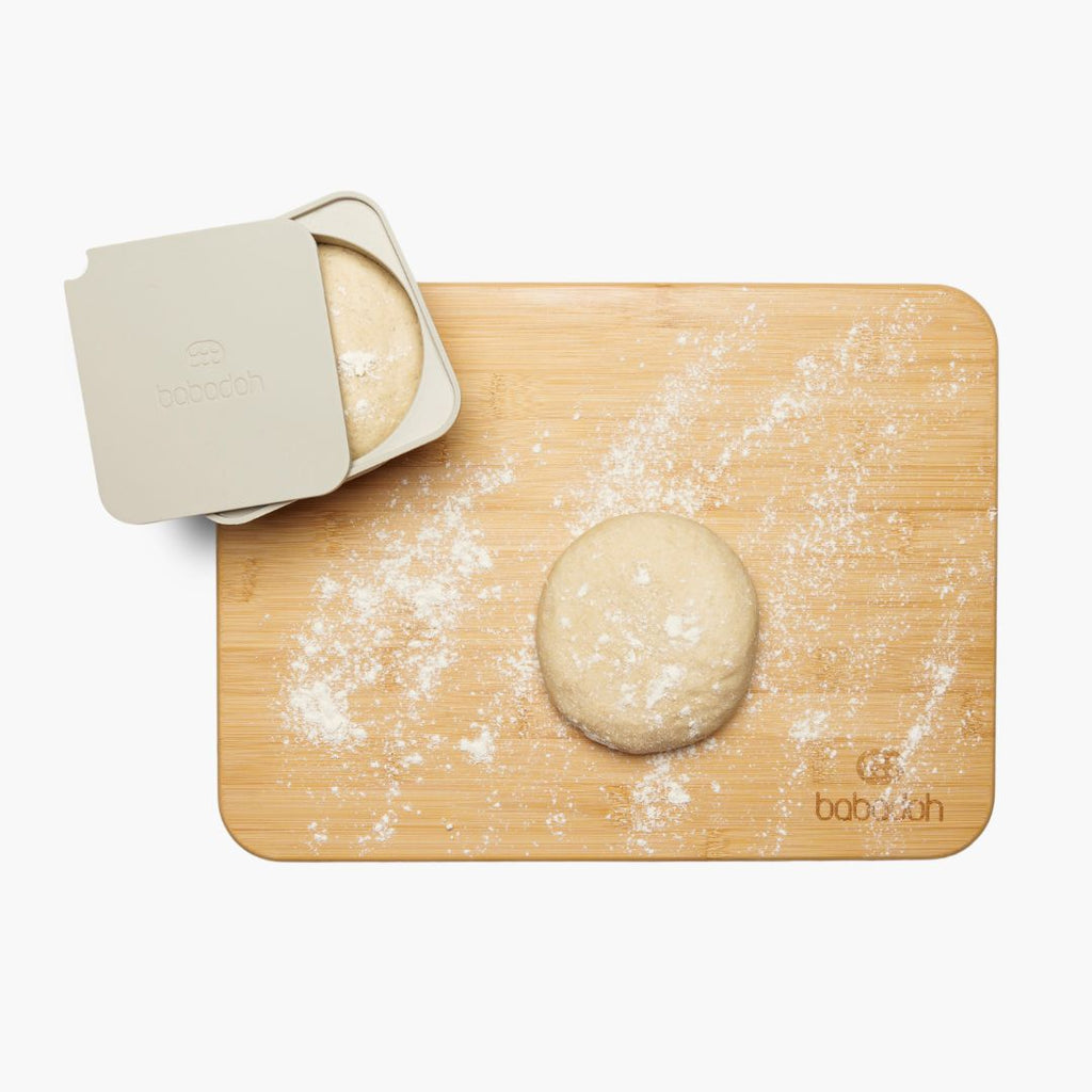 A top-down view of a bamboo cutting board with a round ball of dough resting on it, lightly dusted with flour. A beige container with the brand name 'Babadoh' embossed on the lid holds another ball of dough, partially uncovered.