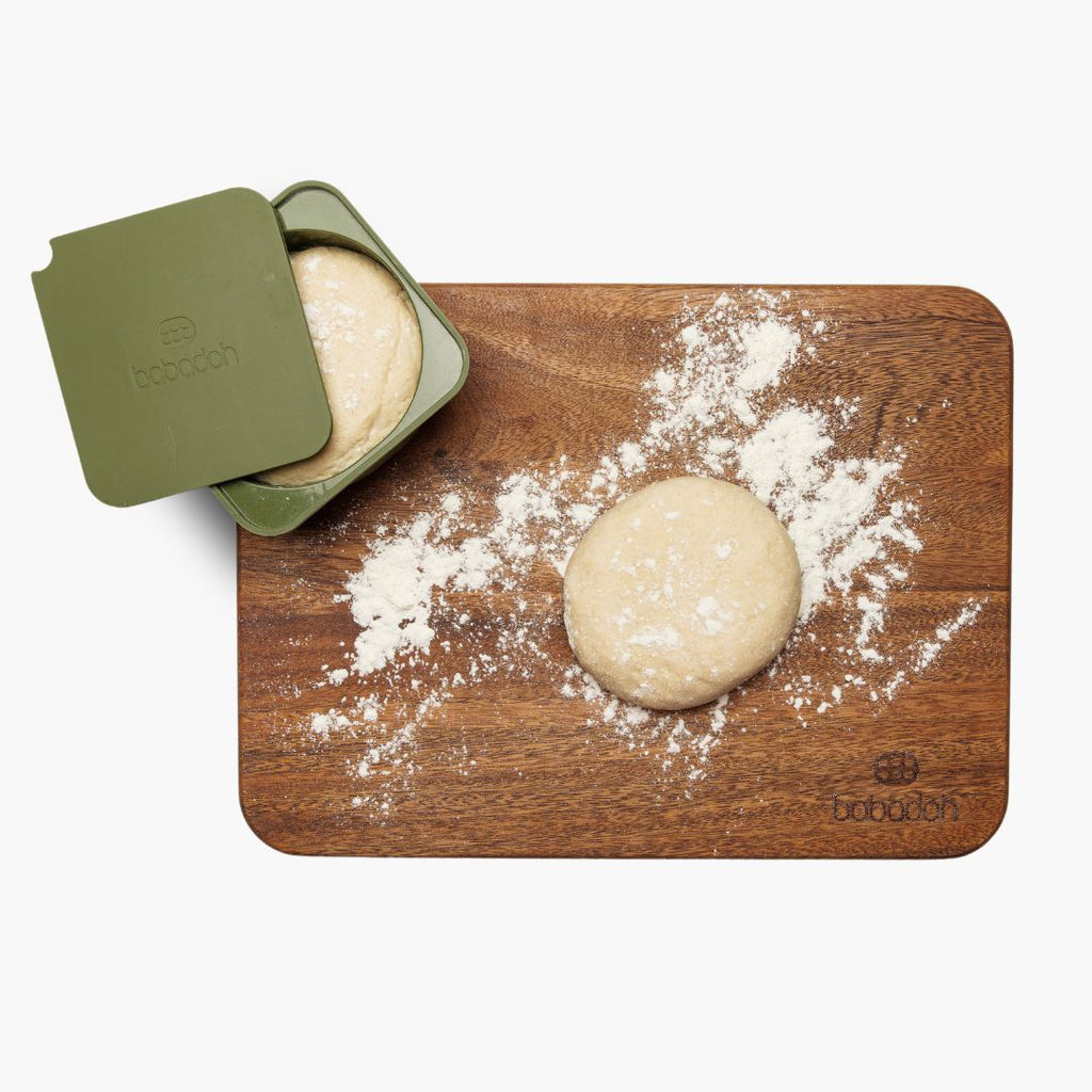 A top-down view of a walnut cutting board with a round ball of dough resting on it, lightly dusted with flour. An olive green container with the brand name 'Babadoh' embossed on the lid holds another ball of dough, partially uncovered.