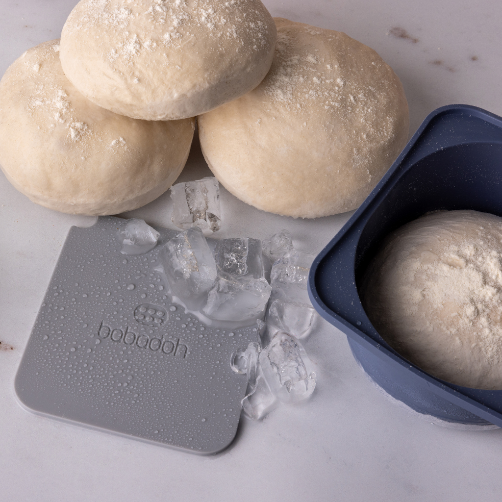 Three frozen dough balls stacked on a marble worktop, with a scattering of ice. A blue container holds another frozen ball of dough.