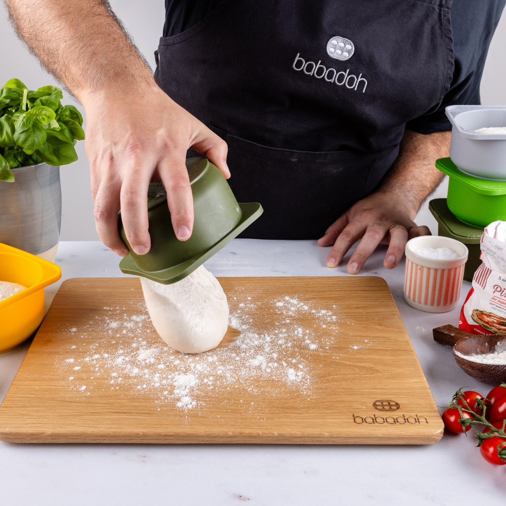 A man wearing a black apron embroidered with 'Babadoh' is standing at a worktop. He is holding an olive green container and dropping a pizza dough ball on to an oak board that is dusted with flour. A basil plant is to his left.
