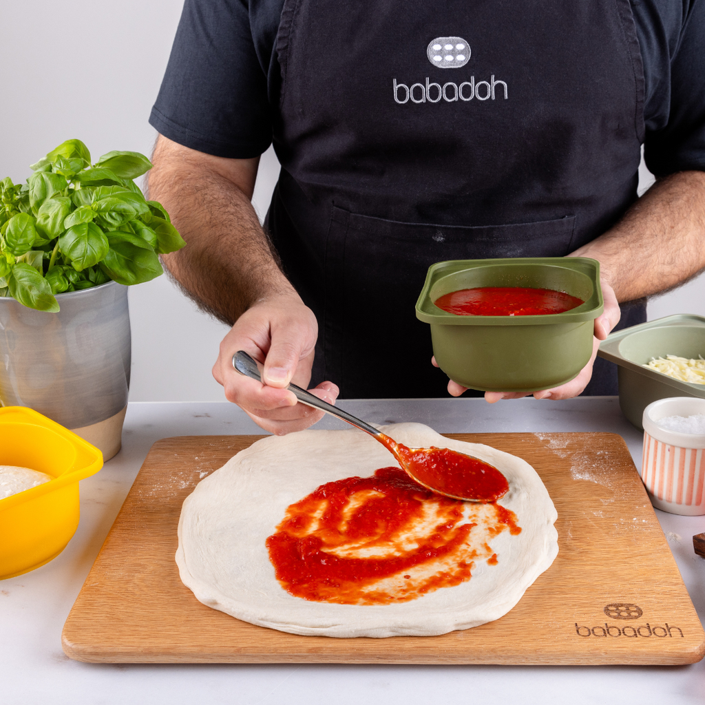 A man wearing a black apron embroidered with 'Babadoh' is standing at a worktop. He is holding on olive green container of pizza sauce and is spreading this across a pizza base.