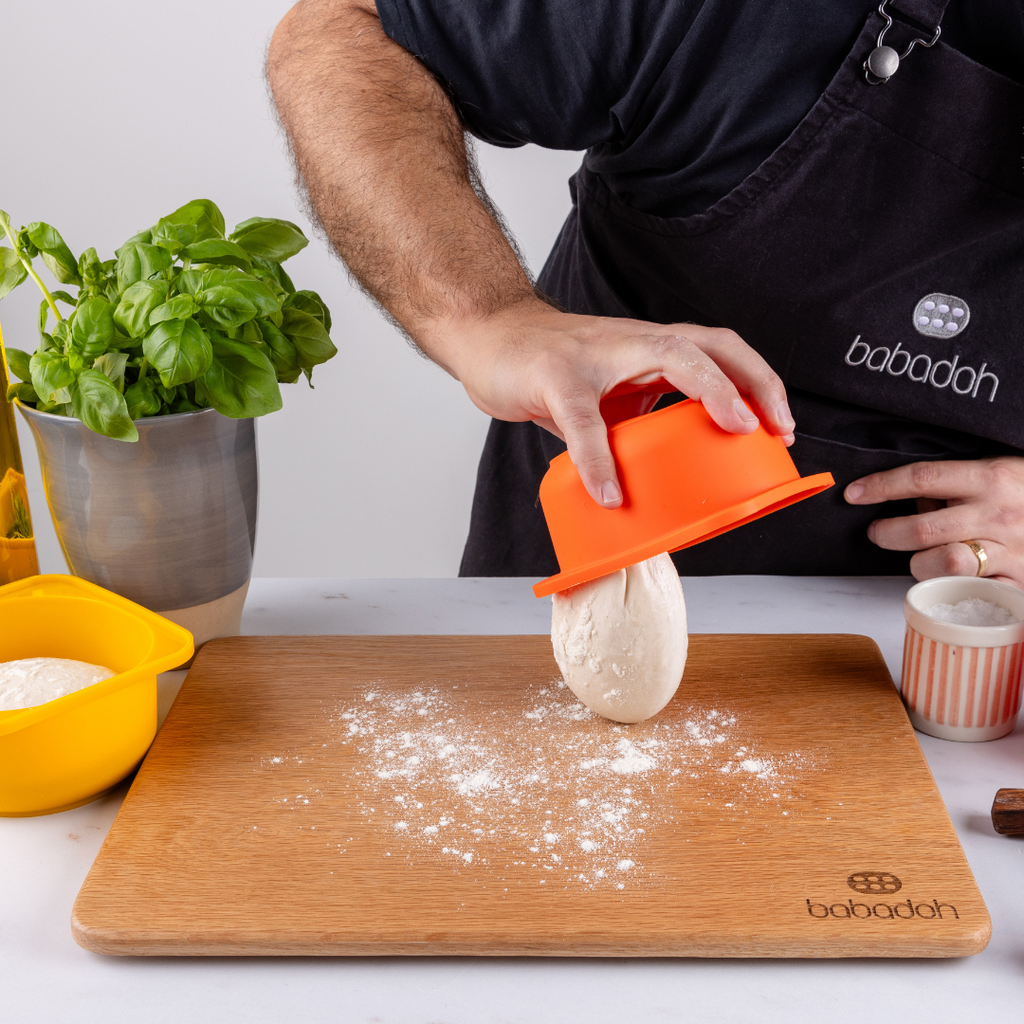 A man wearing a black apron embroidered with 'Babadoh' is standing at a worktop. He is holding an orange container and dropping a pizza dough ball on to an oak board that is dusted with flour. A basil plant and a bottle of olive oil is to his left.