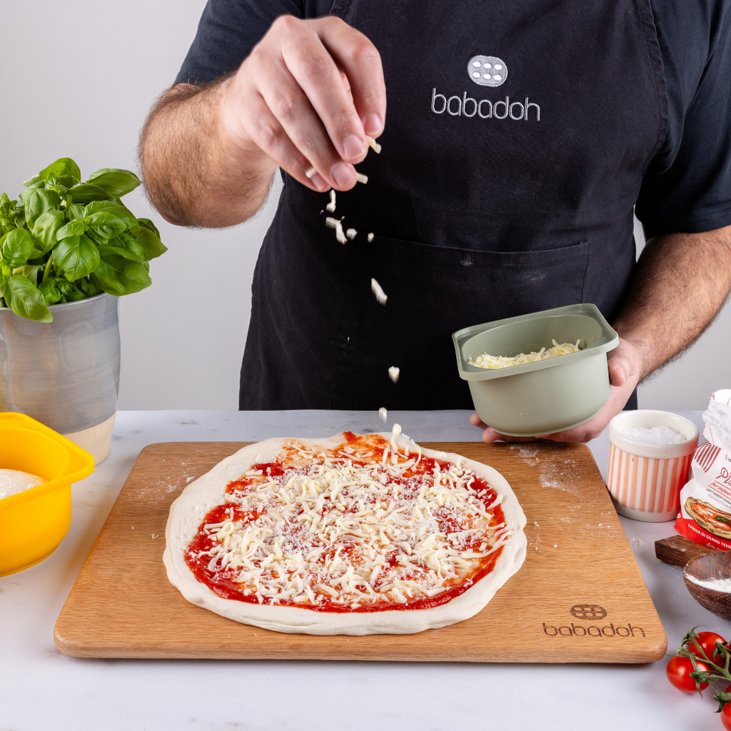 A man wearing a black apron embroidered with 'Babadoh' is standing at a worktop. He is holding a pale green container and scattering grated cheese on to a pizza.