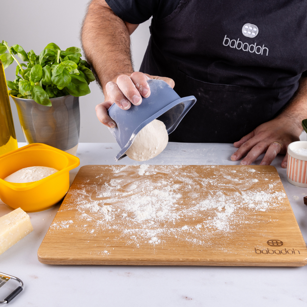 A man wearing a black apron embroidered with 'Babadoh' is standing at a worktop. He is holding a blue container and dropping a pizza dough ball on to an oak board that is dusted with flour. A basil plant and a bottle of olive oil is to his left.
