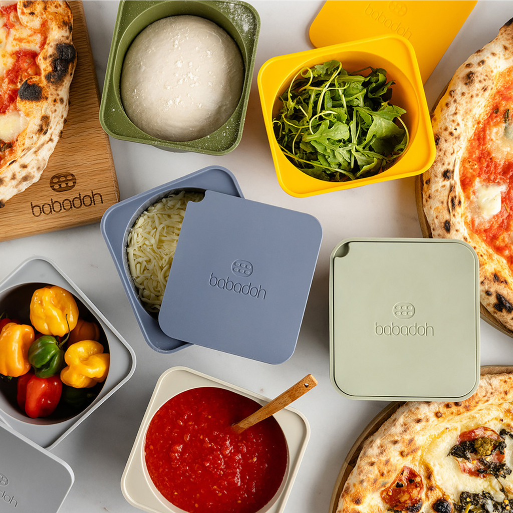 A collection of brightly coloured containers with the brand 'Babadoh', holding a variety of ingredients - red and yellow peppers, grated cheese, rocket, pizza sauce. A trio of pizzas sits around the containers.